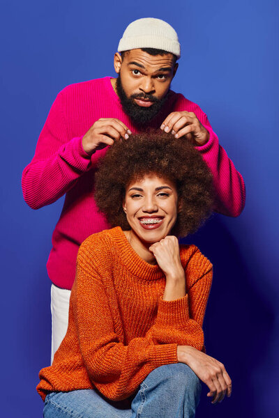 A young African American man and woman sit together, showcasing vibrant casual attire and a strong friendship bond on a blue background.