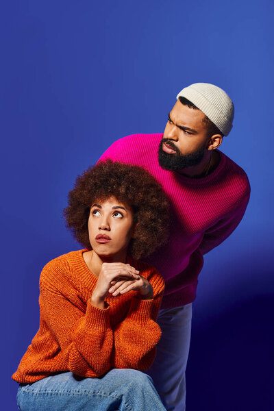 A man and woman, friends, strike a pose in vibrant casual attire against a blue background.