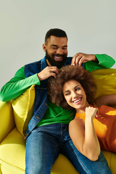 Happy African American man and woman in colorful clothes sitting on a yellow couch against a grey background, symbolizing friendship.