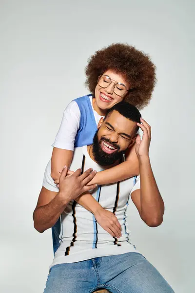 Stock image An African American man in stylish clothes carries a woman on his back against a grey background, showcasing friendship.