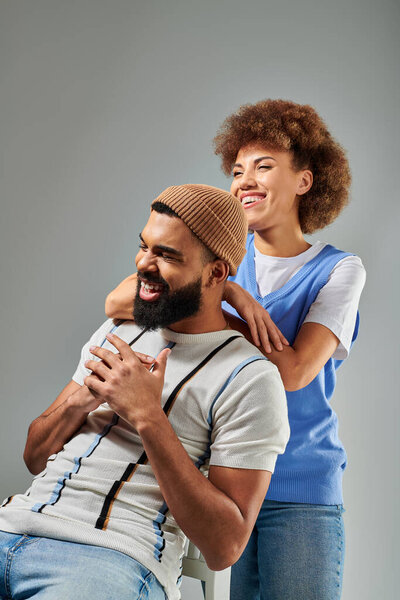 A stylish African American man sitting on top of a chair next to a woman against a grey background, showcasing a unique friendship.