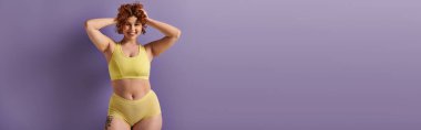 Curvy redhead woman in yellow bikini striking a pose against a vibrant purple backdrop. clipart