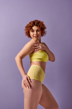 Curvy redhead in yellow underwear poses against a vivid purple backdrop.