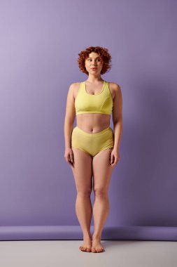 A young, curvy redhead woman in a yellow bikini stands confidently in front of a vibrant purple wall.