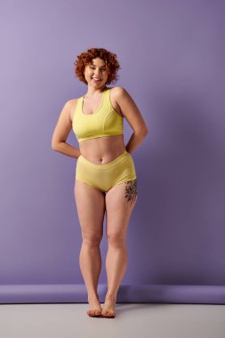 Curvy redhead woman flaunting a yellow bikini in front of a vibrant purple wall. clipart