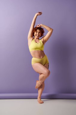 Young, curvy redhead woman in a yellow bikini striking a confident pose in front of a vibrant purple wall.