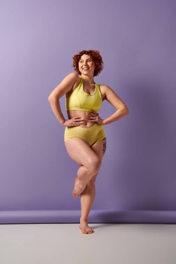 Curvy redhead beauty in yellow bikini striking a pose in front of a vibrant purple wall.