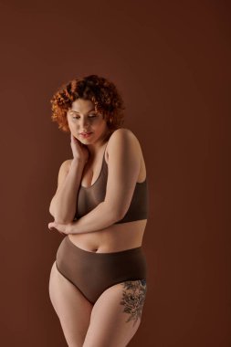 A curvy redhead woman strikes a pose in a brown bikini against a matching background.