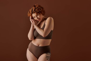 Young, curvy redhead woman exudes confidence in brown bikini on brown background.