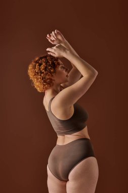 Young curvy redhead woman in brown bikini striking a pose with hands raised.