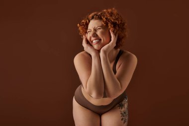 A young, curvy redhead woman poses seductively in a bikini against a warm brown backdrop.