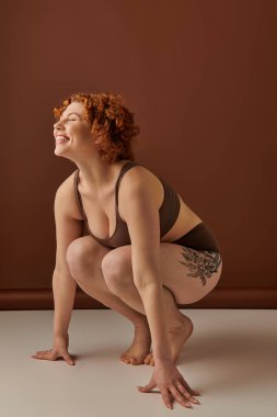 Young, curvy redhead woman crouches gracefully in brown underwear on a textured earth-toned background.