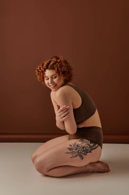 A young, curvy redhead woman sits gracefully on the floor in a cozy brown room. clipart