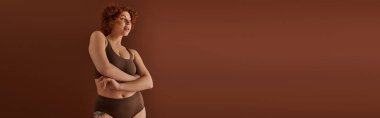 A young, curvy redhead woman in a bikini exudes confidence in front of a brown background.