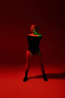 A young, curvy redhead woman stands confidently in front of a vibrant red background.