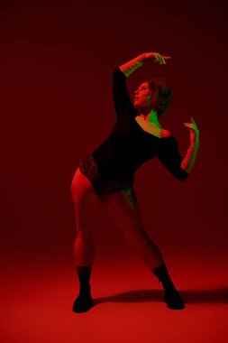A young curvy redhead woman in a bodysuit dances gracefully in front of a vivid red background.