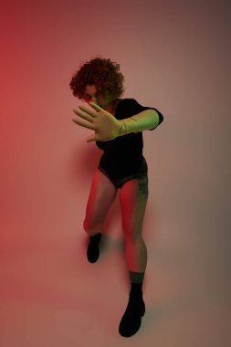 A young curvy redhead woman in a bodysuit dances energetically in front of a red spotlight.