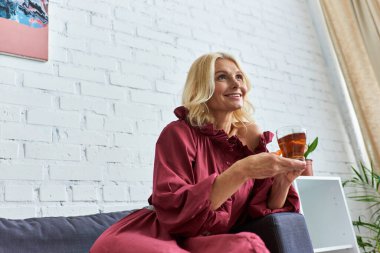 A mature woman in a stylish dress relaxes on a couch, holding a cup of tea. clipart