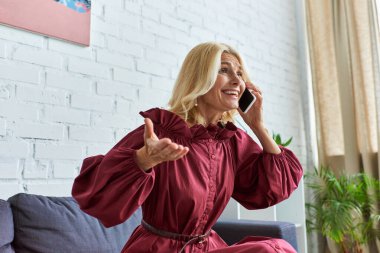 Geniş elbiseli şık bir kadın koltukta otururken cep telefonuyla konuşuyor..