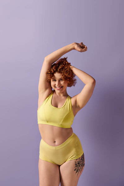 A young, curvy redhead woman in a yellow bikini poses confidently on a purple background.