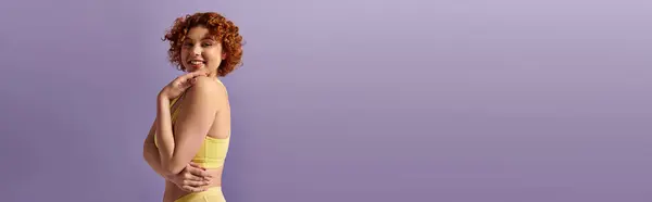 stock image A young, curvy redhead woman poses beautifully in a yellow underwear against a vibrant purple backdrop.