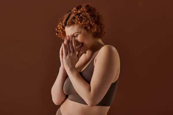 stock image Curvy redhead woman in brown bikini, hands covering face delicately.