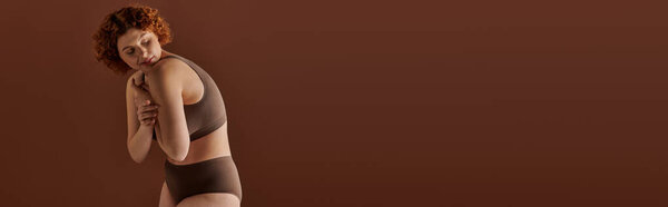 A voluptuous redhead woman poses in a bikini against a rich brown backdrop.