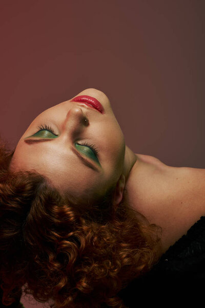 A young, curvy redhead woman in a bodysuit is laying down, exuding an ethereal aura under soft red lighting.