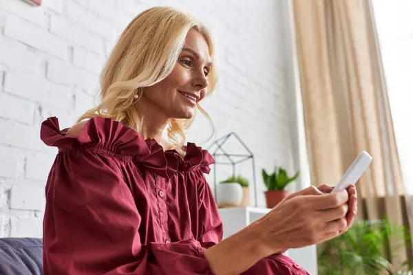 stock image Mature woman in a chic dress, sitting on a couch, captivated by her cell phone.