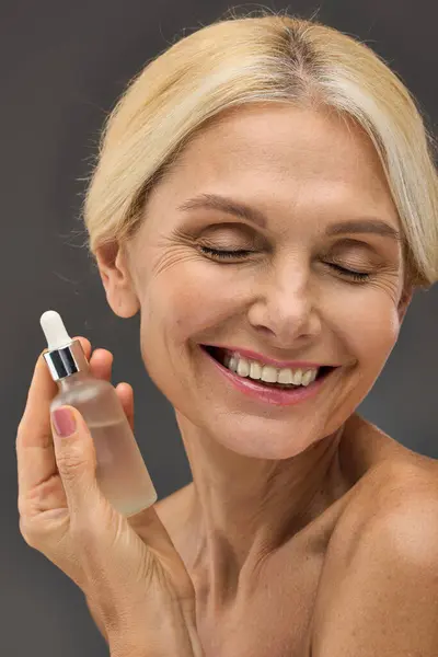 stock image Beautiful mature woman holding serum on a gray backdrop.