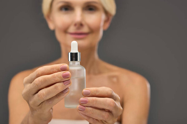Attractive mature woman holding serum on a gray backdrop.
