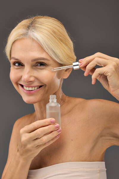 Good looking mature woman applying serum on a gray backdrop.