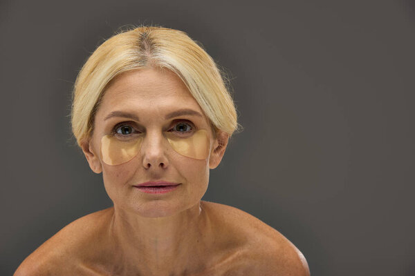 Довольно зрелая женщина с светлыми волосами с повязками на глазах на сером фоне.