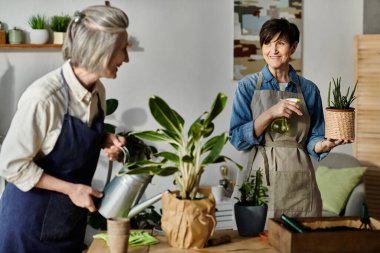 Two women in aprons tend to potted plants in a kitchen. clipart