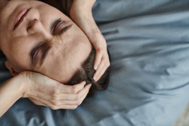 Middle-aged woman in bed, hands on head, deep in thought.