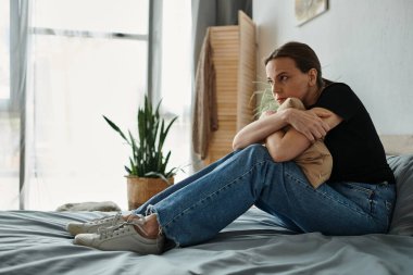 A middle-aged woman in jeans and a t-shirt sits peacefully on a bed at home.