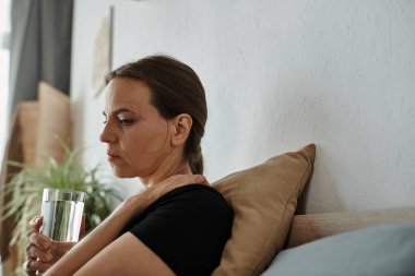 Middle-aged woman sitting on a bed holding a glass of water.