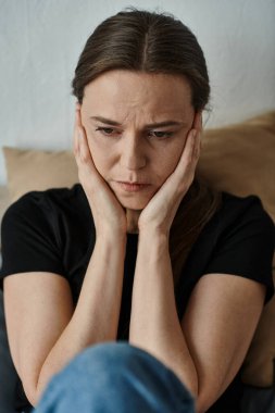 Middle-aged woman with hands on face sitting on couch at home.