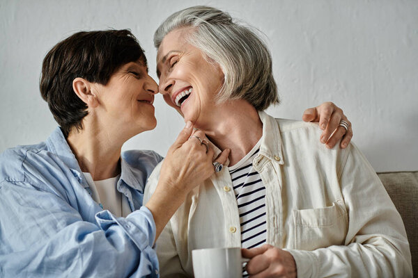 Two older women, a loving mature lesbian couple, share a cup of coffee on a cozy couch.