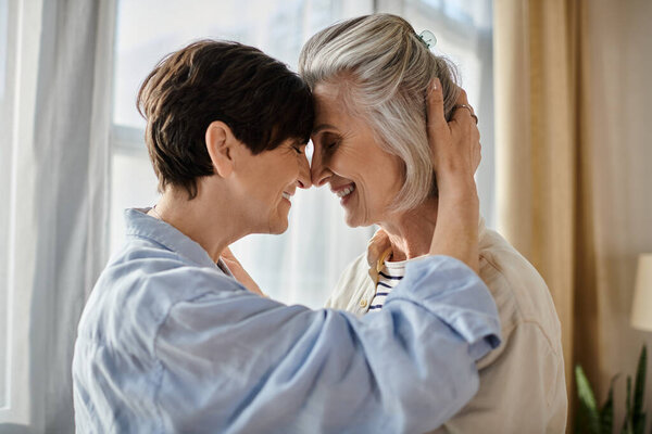 Two older women embrace lovingly in front of a window.