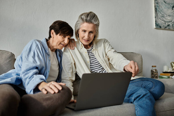 Two older women, a loving mature lesbian couple, sit on a couch, engrossed in a laptop.