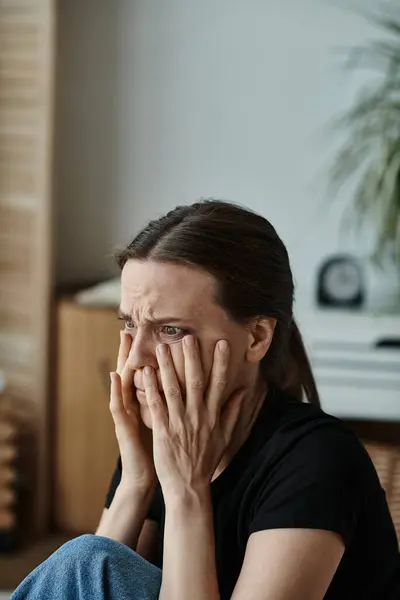 stock image A middle-aged woman sits at home, covering her face with her hands in distress.