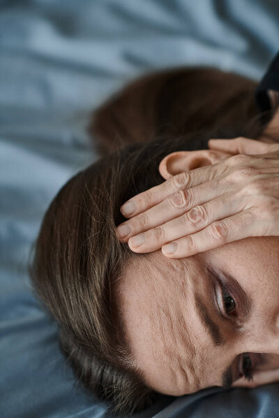 A middle-aged woman laying on a bed, holding her head in distress.