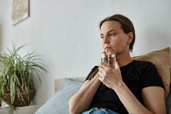 A middle-aged woman sits on a bed holding a glass of water, lost in thought.