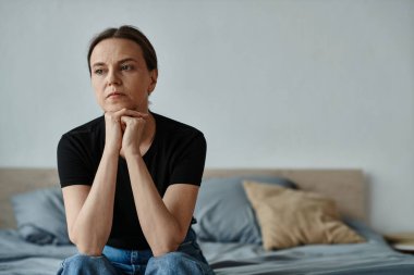 Middle-aged woman sitting on bed, deep in thought, hand on chin.