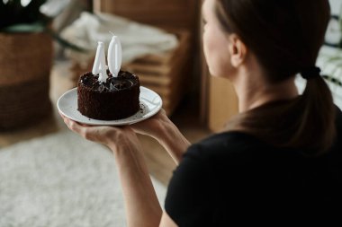A middle-aged woman holding a birthday cake on a plate in a home setting.