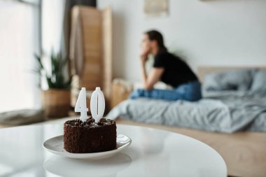 A middle-aged woman sits alone on a bed with a cake, lost in thought.