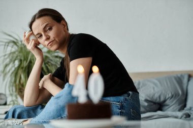 Middle-aged woman in deep thought sits with birthday cake in front of her.