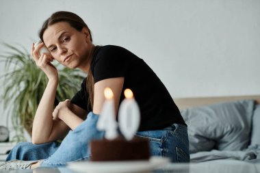 Middle-aged woman in deep thought sits with birthday cake in front of her. clipart
