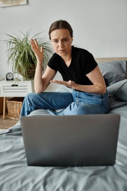 Woman sits on bed with laptop, seeking solace in virtual therapy. clipart